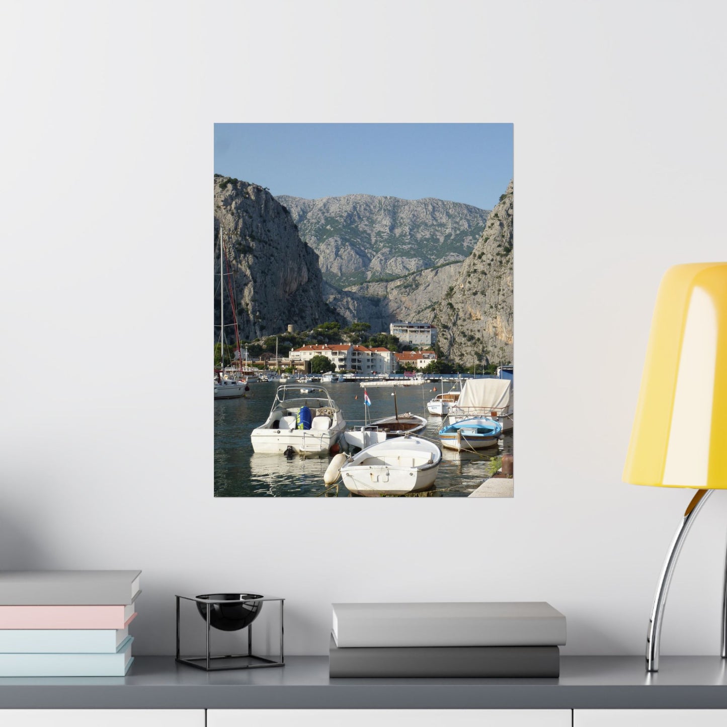 Framed photograph of a coastal scene with boats and mountains on a wall above a shelf.