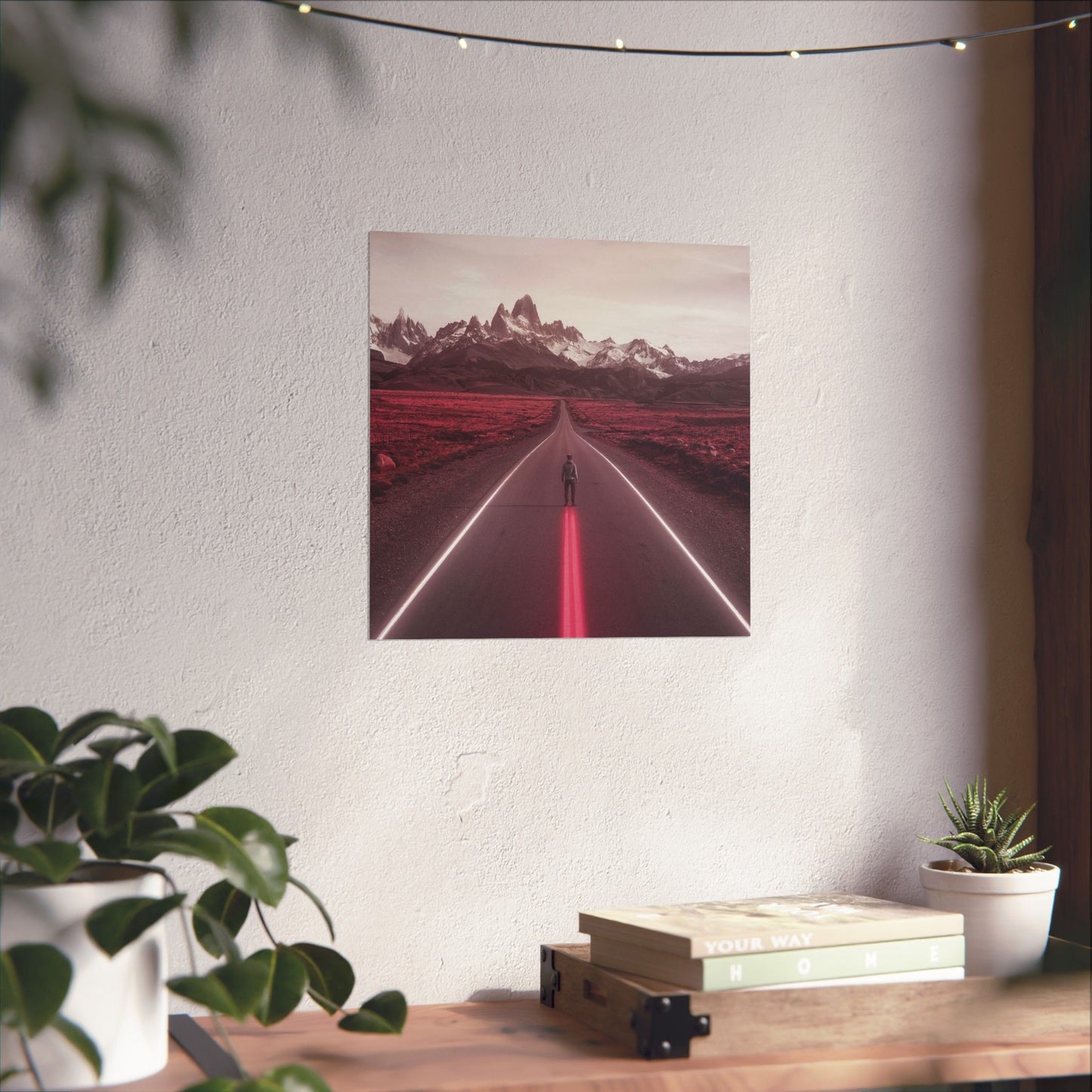 Framed artwork of a road leading to mountains on a white wall above a wooden desk with plants.