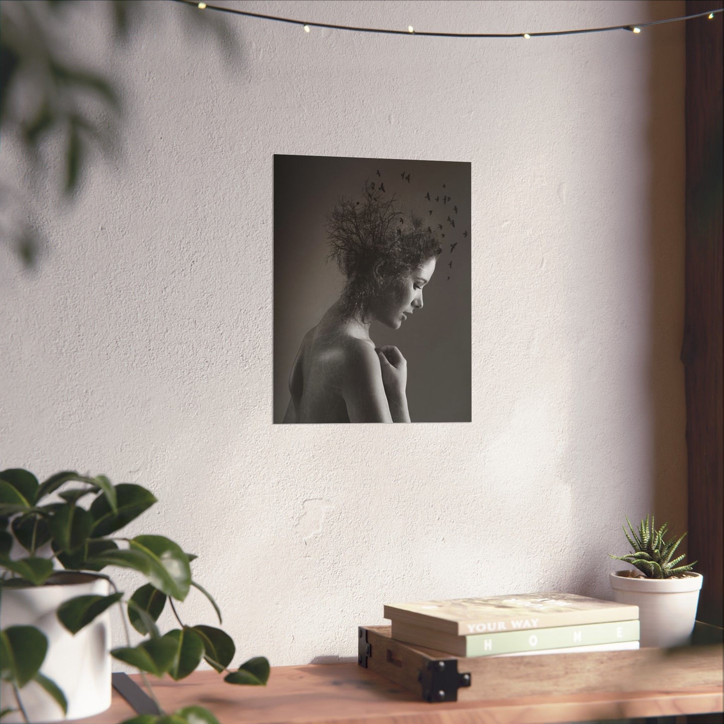 Wall art of a woman with birds on a white wall above a wooden table with plants and books.