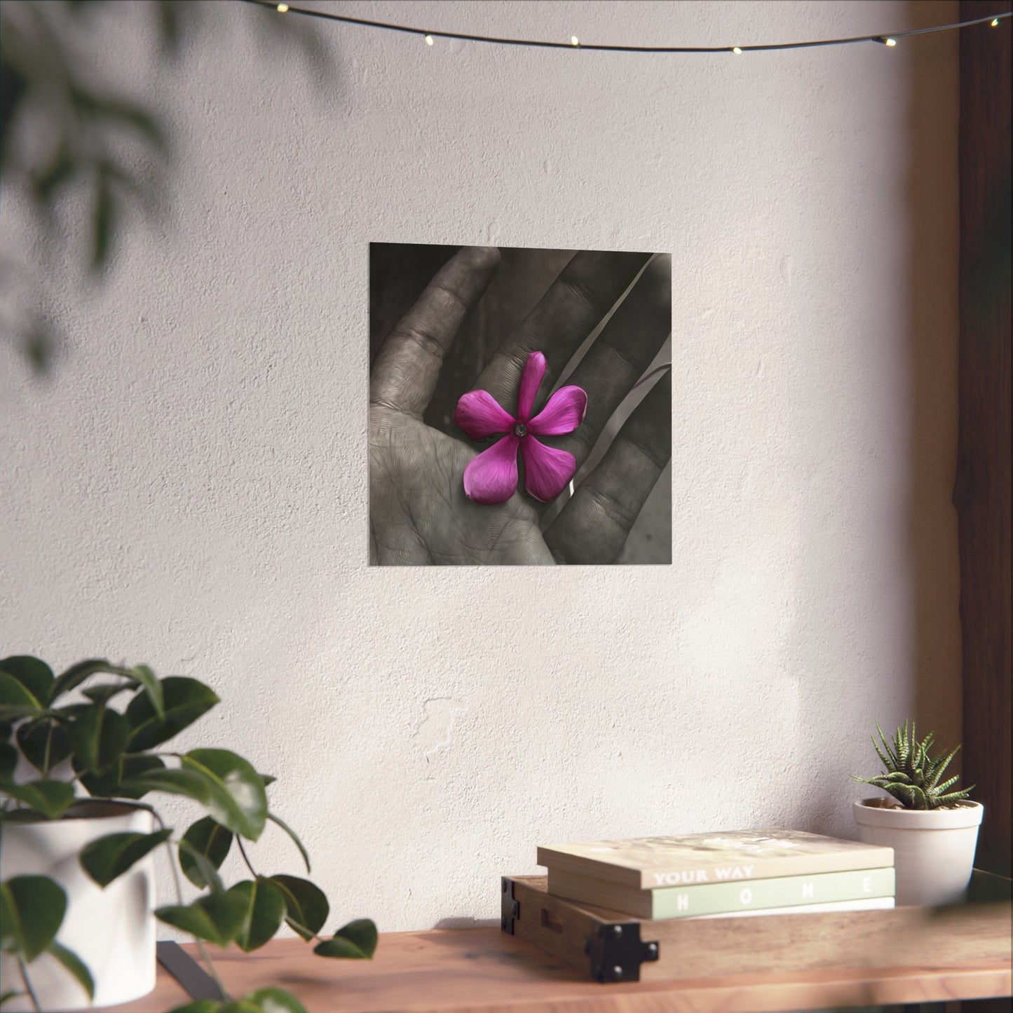 Framed artwork of a pink flower held in a hand on a wall above a wooden table with plants.