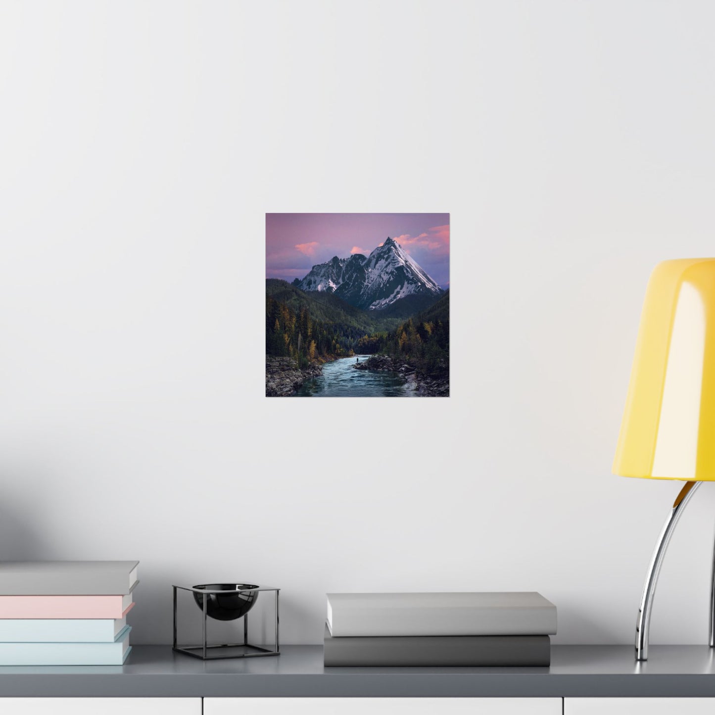 Framed mountain landscape print on a wall above a desk with books and a lamp.