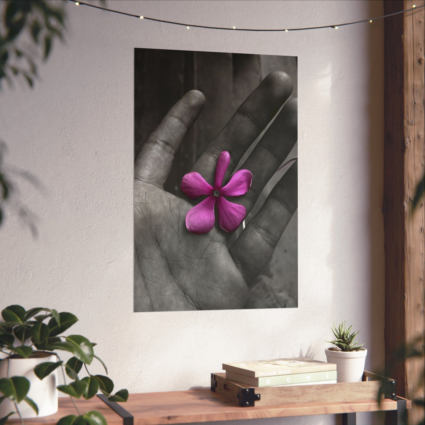 Framed artwork of a hand holding a pink flower on a wall above a wooden shelf with books and plants.