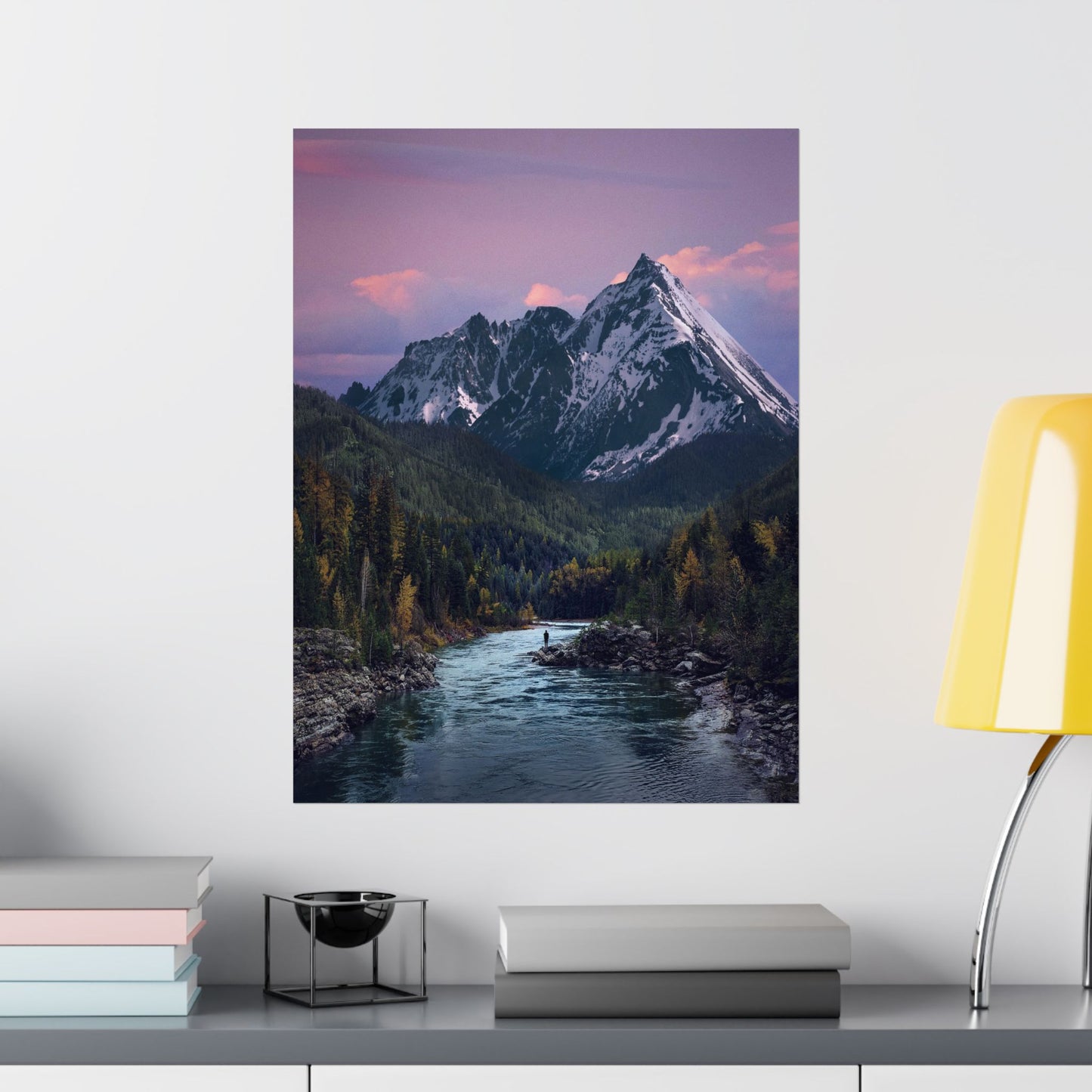 Framed mountain landscape print on a wall above a shelf with books and decor items.