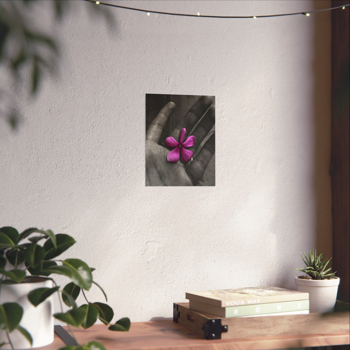 Hand holding a pink flower on a wall above a wooden desk with plants and books.