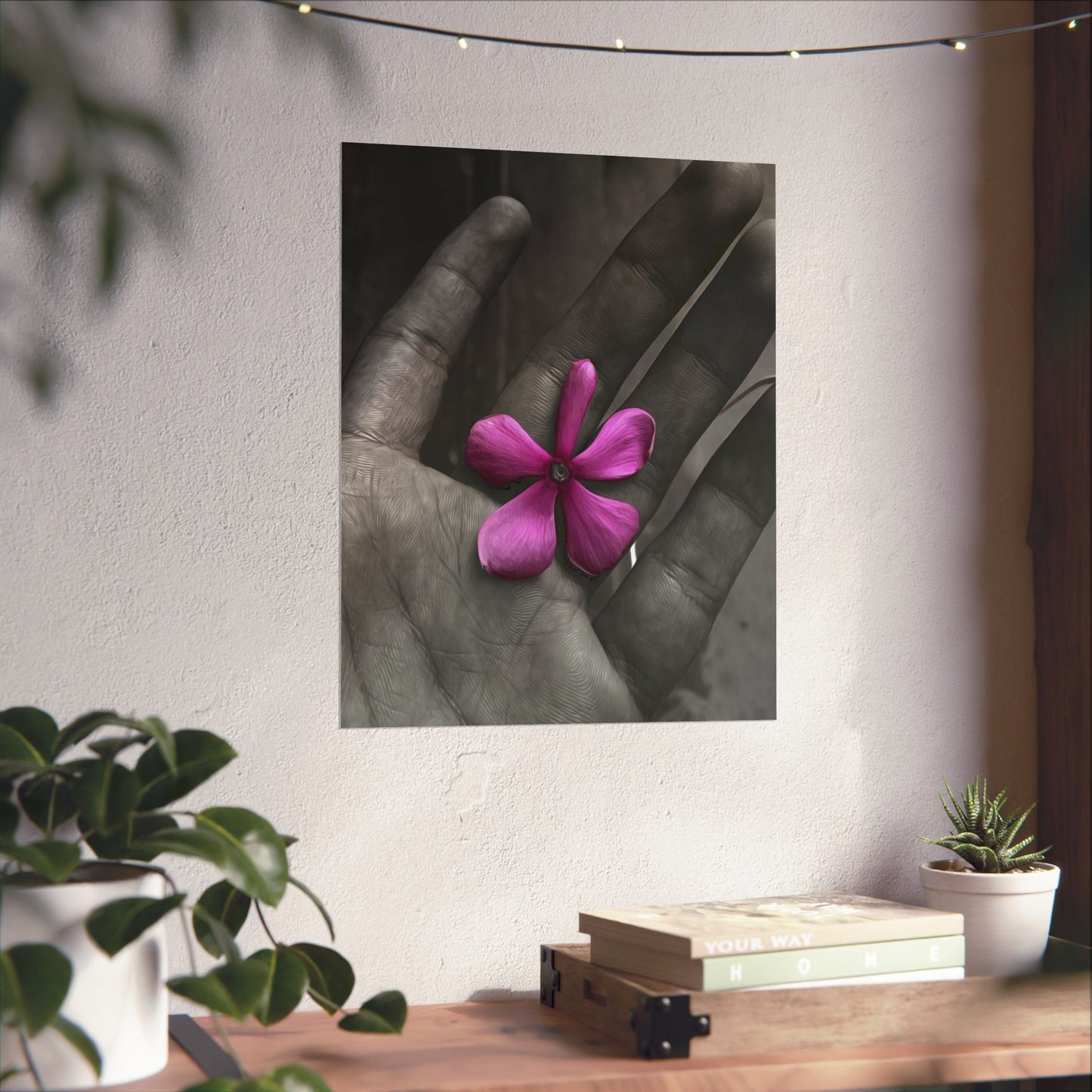 Framed artwork of a hand holding a pink flower on a wall with plants and books below.