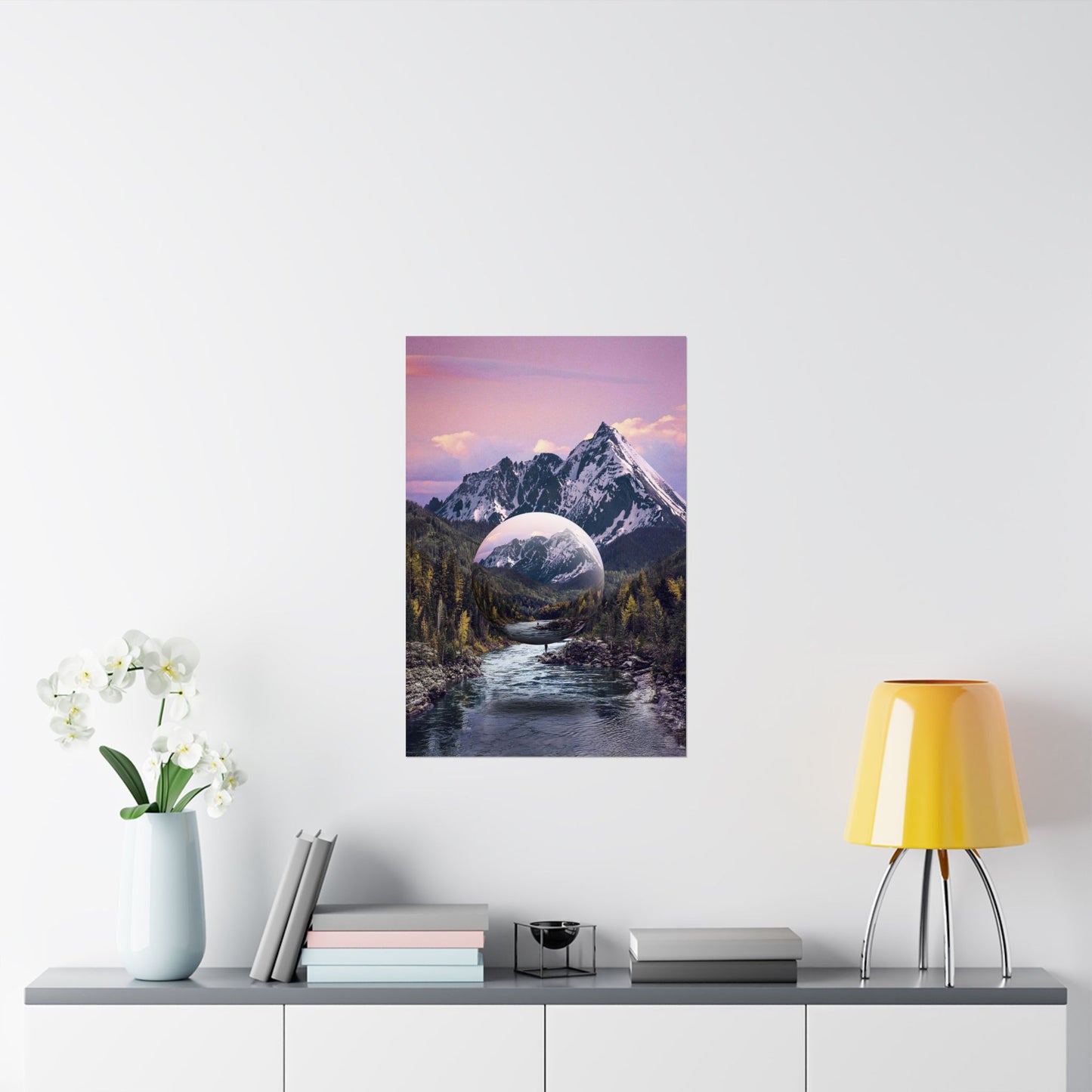 Framed artwork of a mountain landscape with a river and a planet in the sky, displayed on a wall above a shelf.