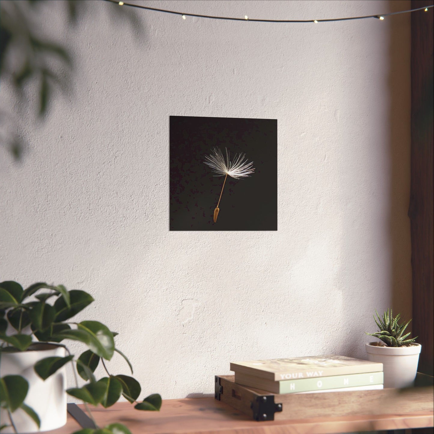Wall art of a dandelion seed head on a plain wall with plants and books on a table below.
