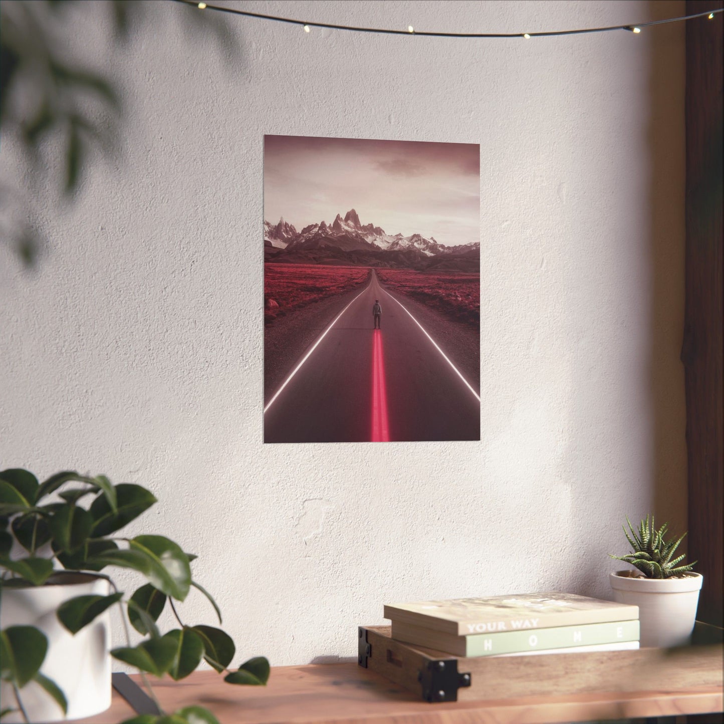Poster of a red road with mountains on a white wall above a wooden desk with plants and books.