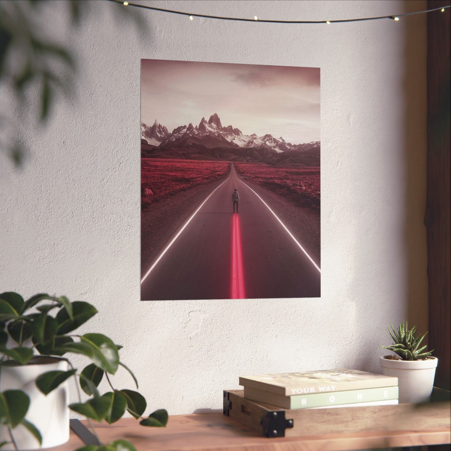 Poster of a red-lit road with mountains on a white wall above a desk with plants and books.