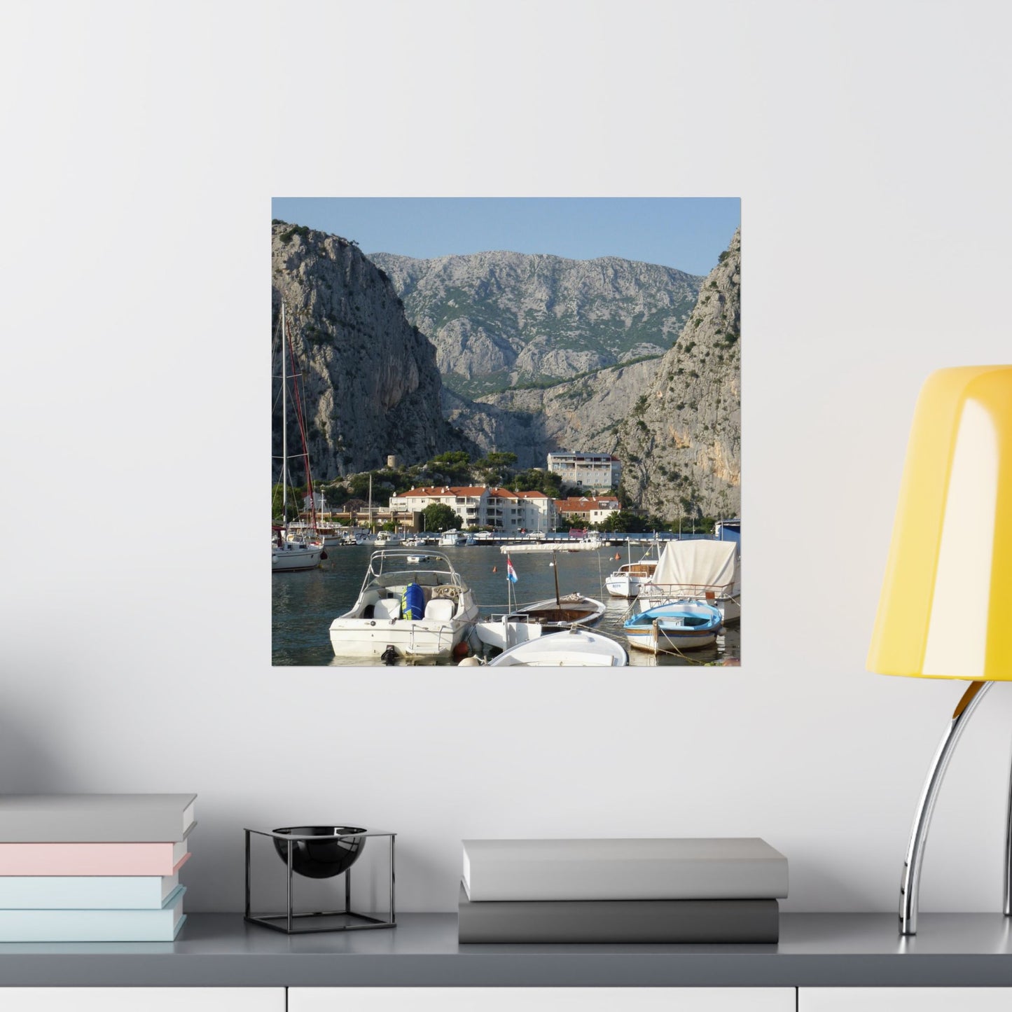 Canvas print of a coastal scene with boats and mountains on a wall above a shelf.