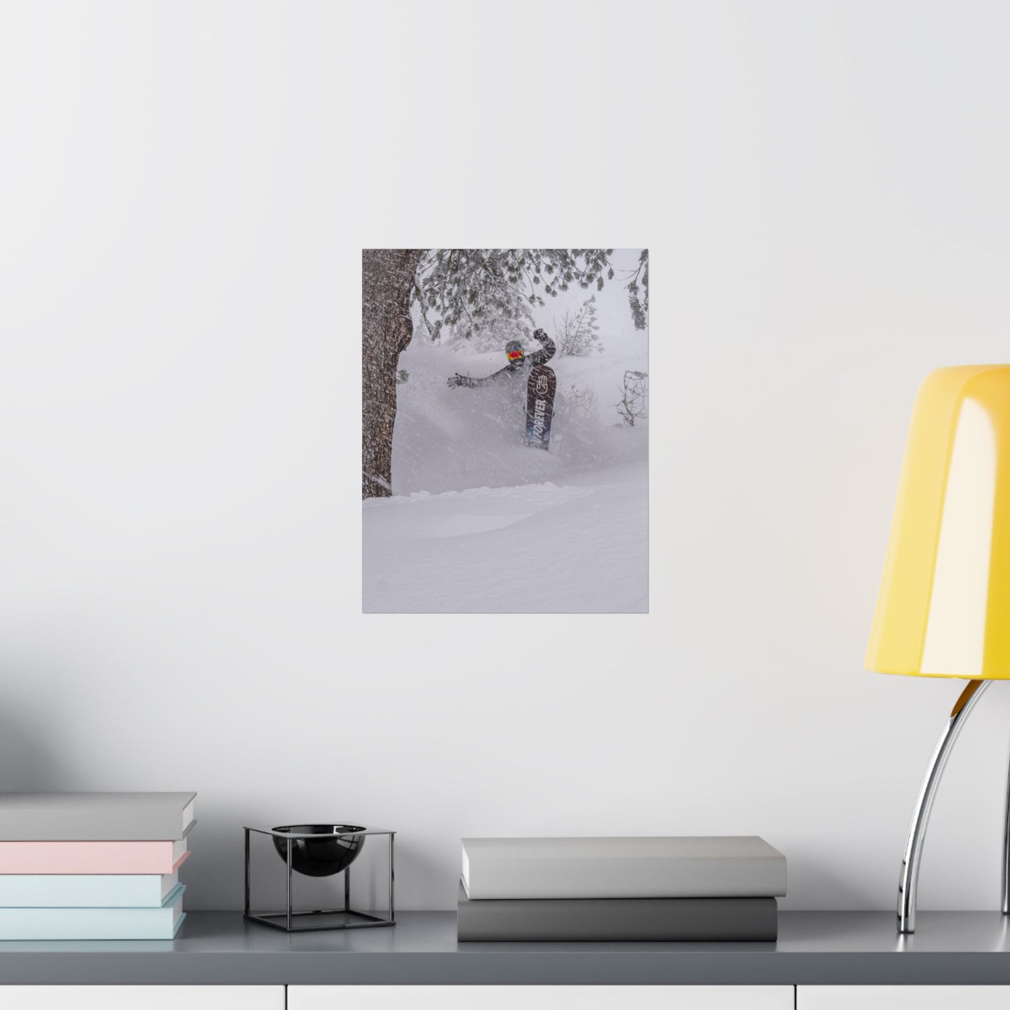 Poster of a skier in a snowy forest on a white wall above a desk with books and a lamp.