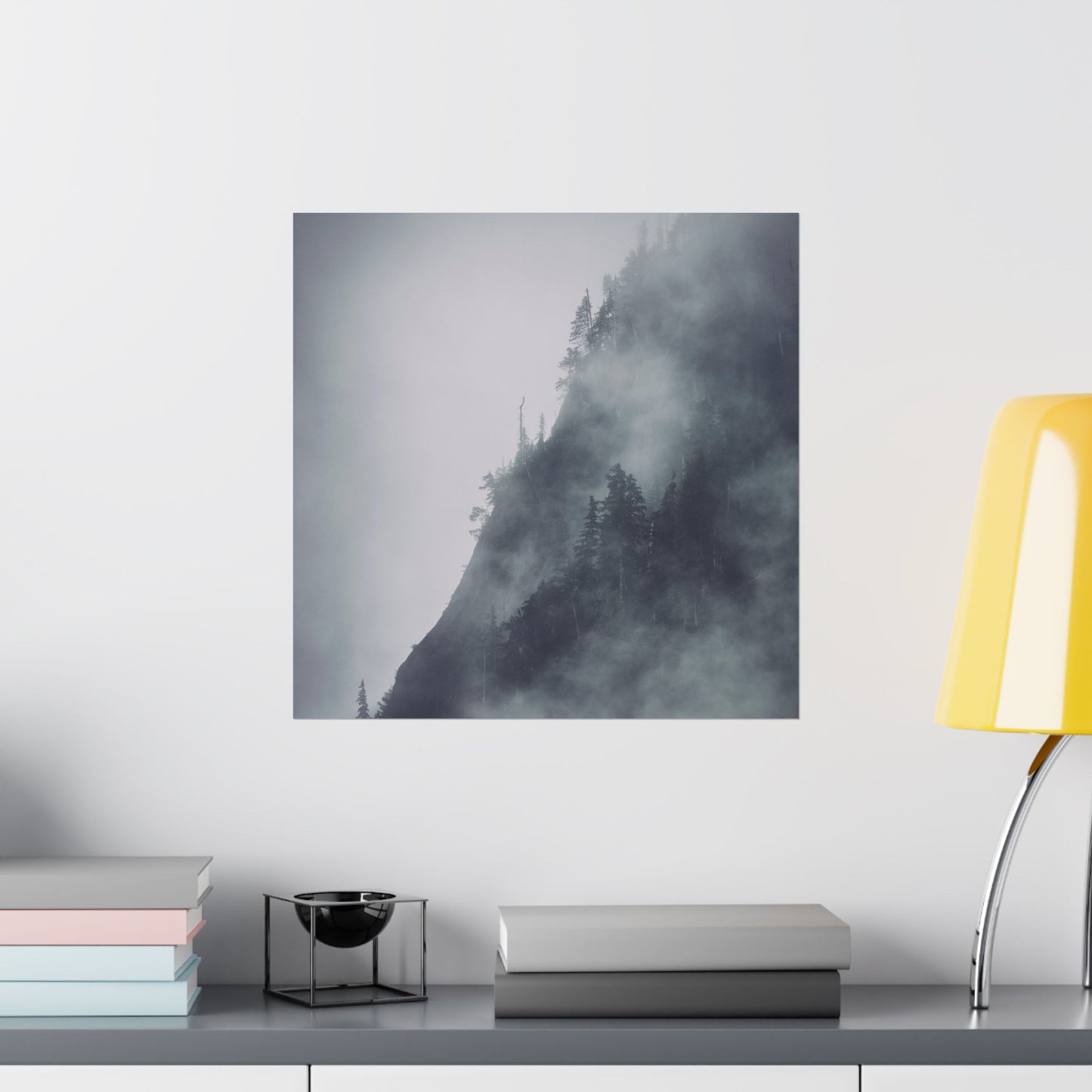 Foggy mountain landscape print on a wall above a shelf with books and a lamp.