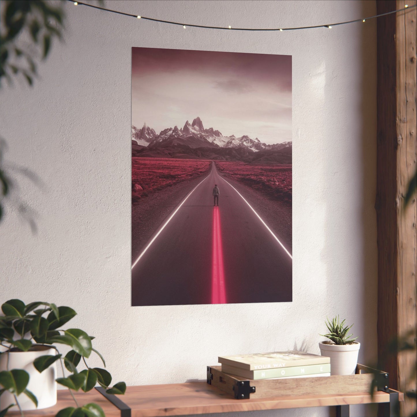 Poster of a road with mountains on a wall above a wooden shelf with books and plants.