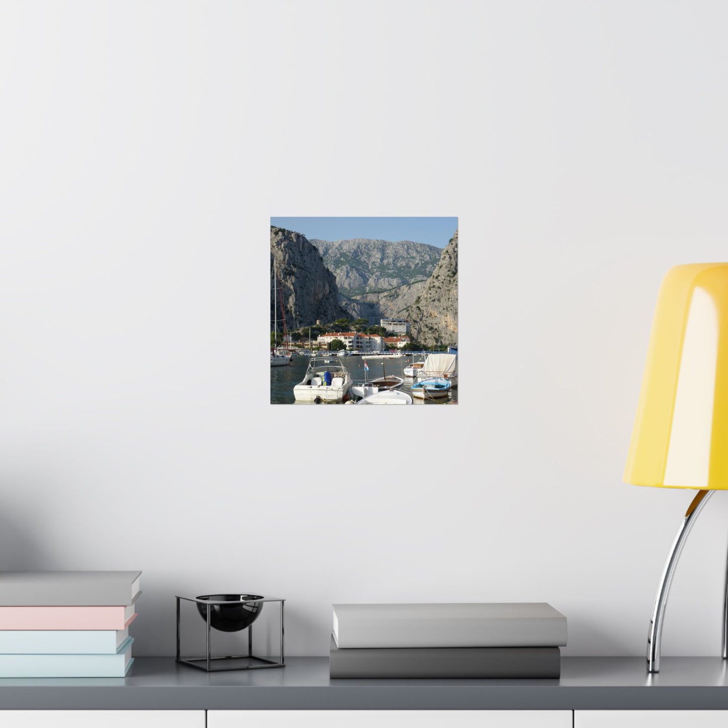 Wall art of a coastal scene with mountains and boats on a white wall above a shelf.