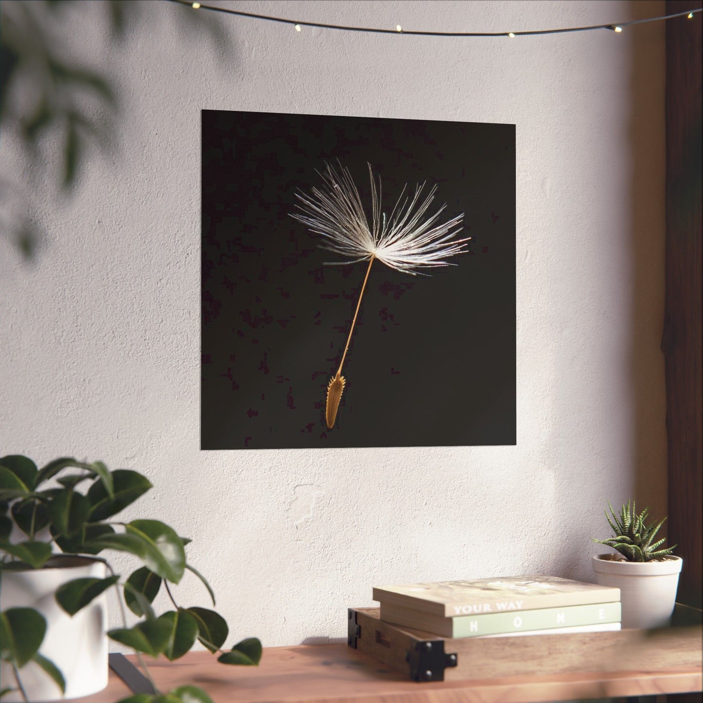 Framed artwork of a dandelion seed on a wall above a wooden table with plants and books.