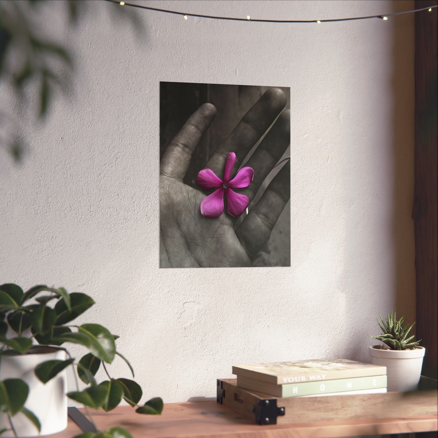 Framed artwork of a hand holding a pink flower on a wall above a wooden table with plants.