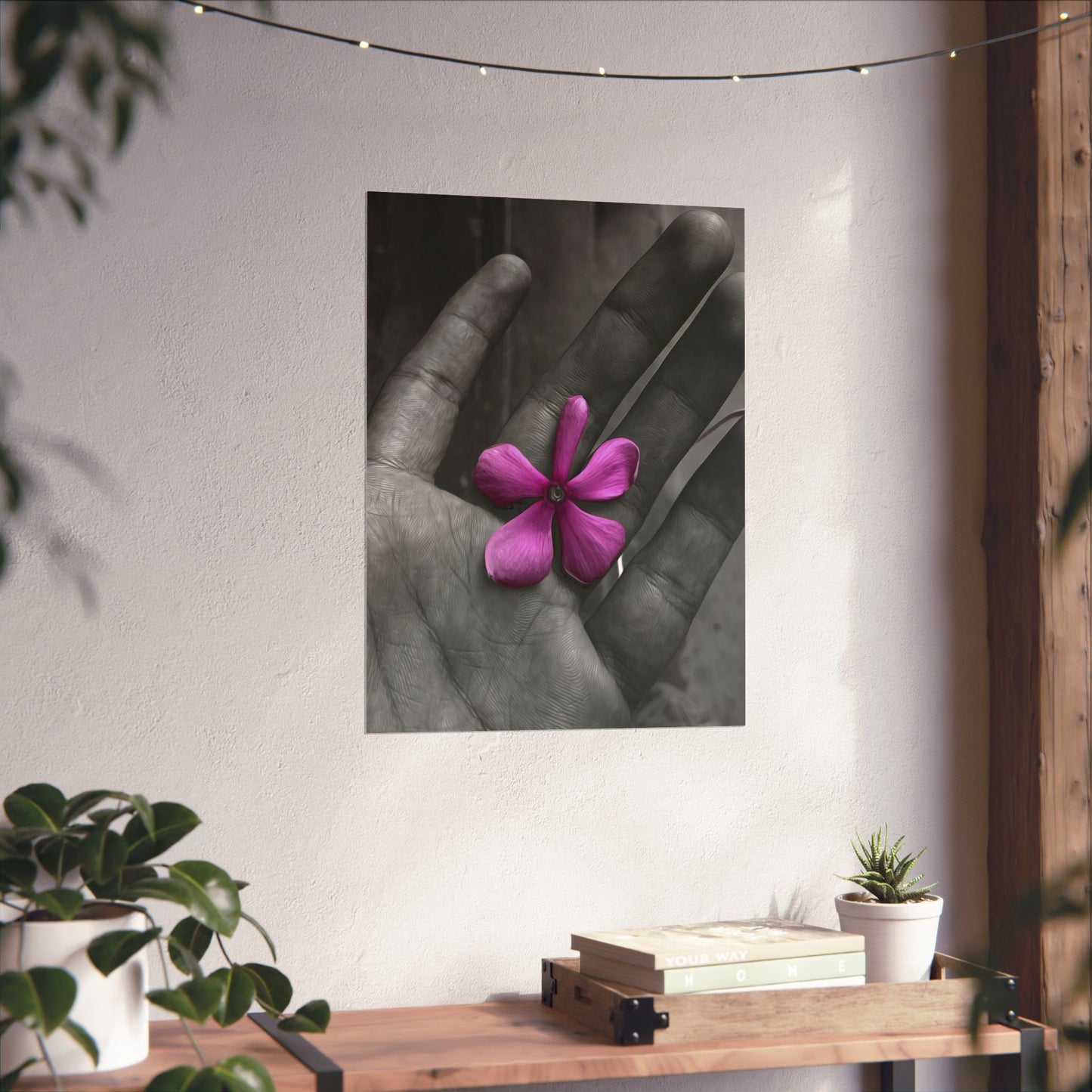 Framed artwork of a hand holding a pink flower on a wall above a wooden shelf with books and plants.