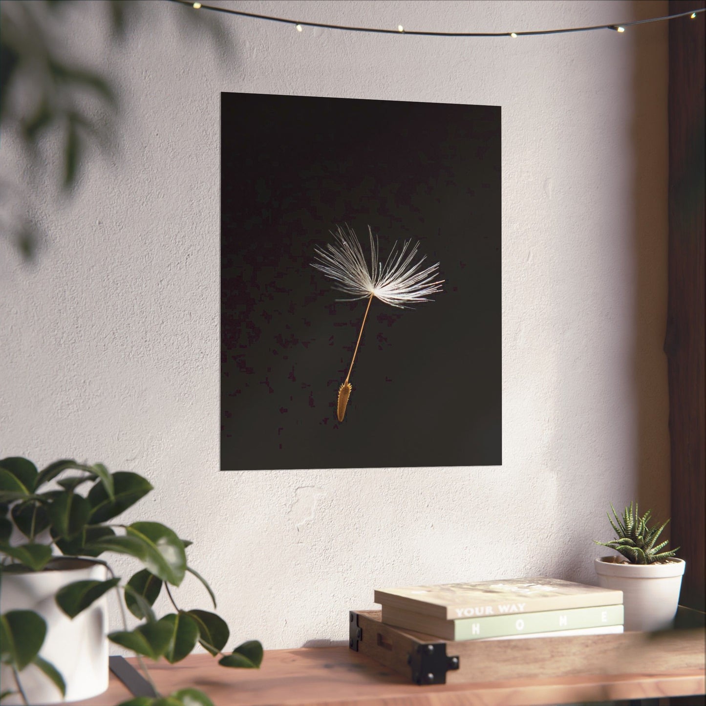 Framed artwork of a dandelion seed on a wall with plants and books below.