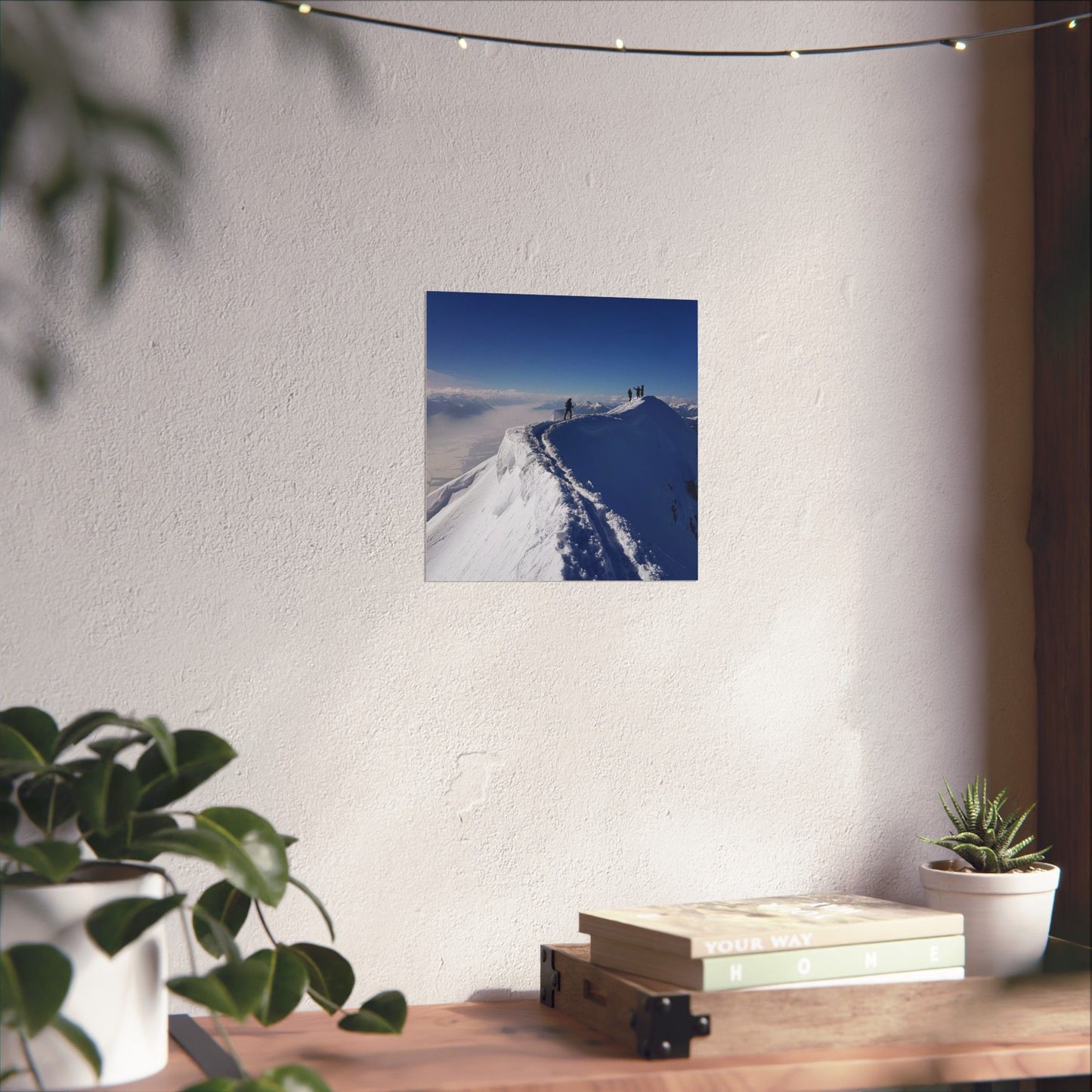 Poster of a snowy landscape with mountains on a white wall above a wooden desk with plants and books.