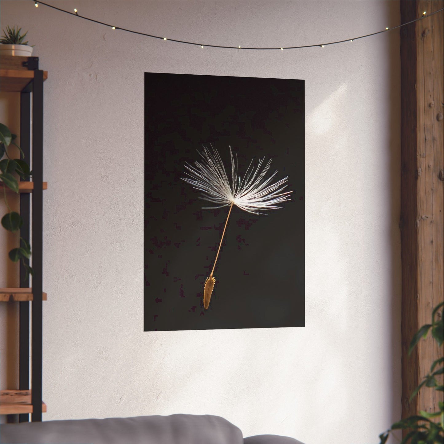 Framed artwork of a dandelion seed head on a dark background in a room with string lights and a plant.