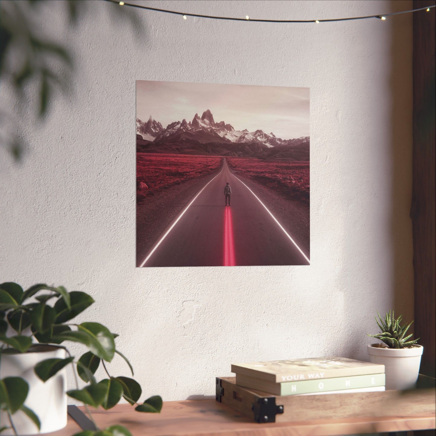 Framed artwork of a road with mountains on a white wall above a desk with plants and books.