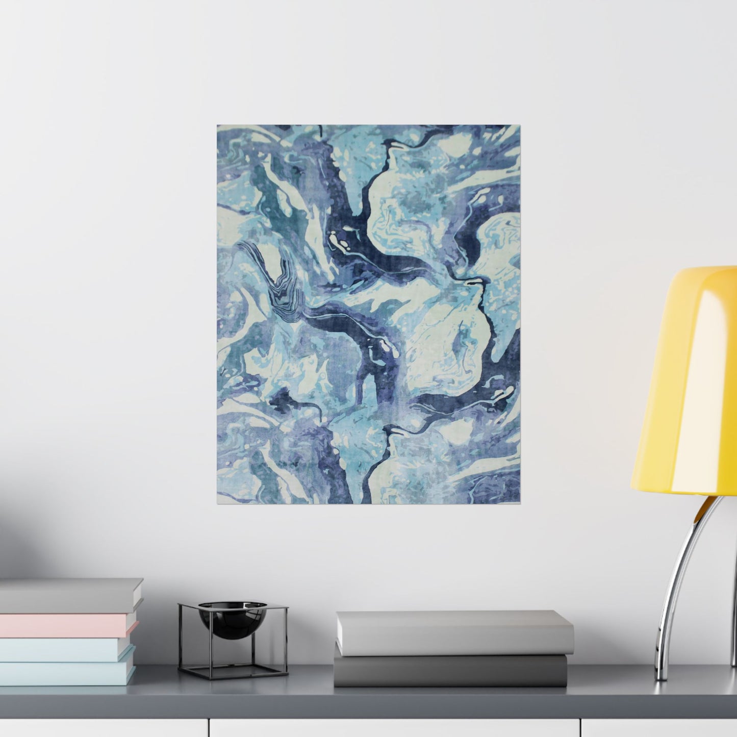 Abstract blue and white painting on a wall above a shelf with books and a lamp.
