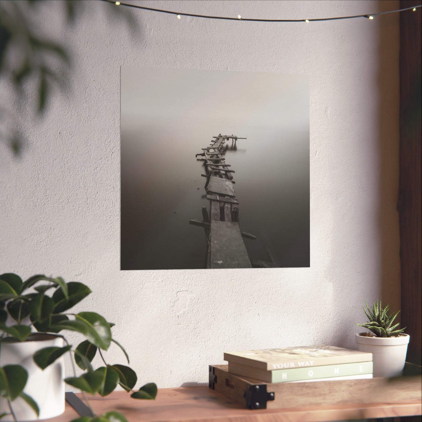 Framed black and white photograph of a dock on a wall above a wooden desk with plants and books.