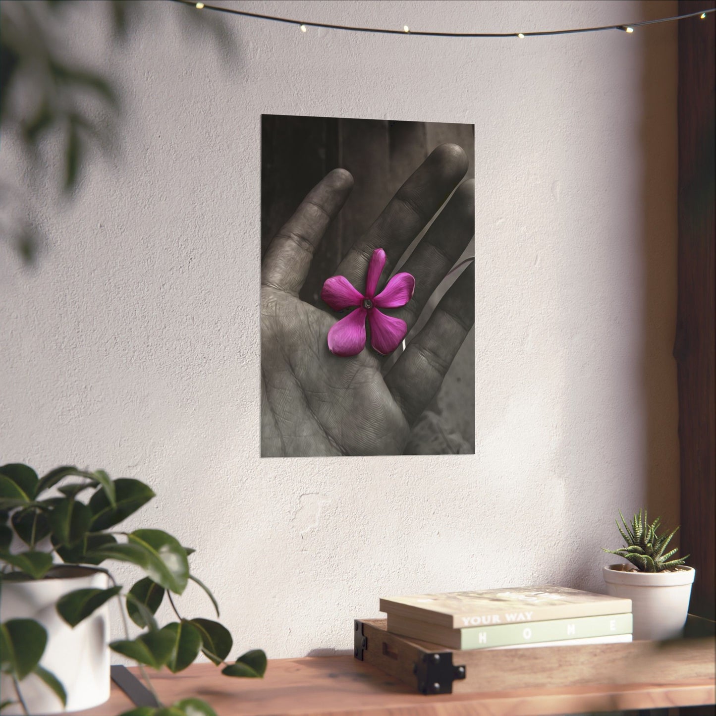 Framed artwork of a hand holding a pink flower on a wall above a wooden desk with plants.