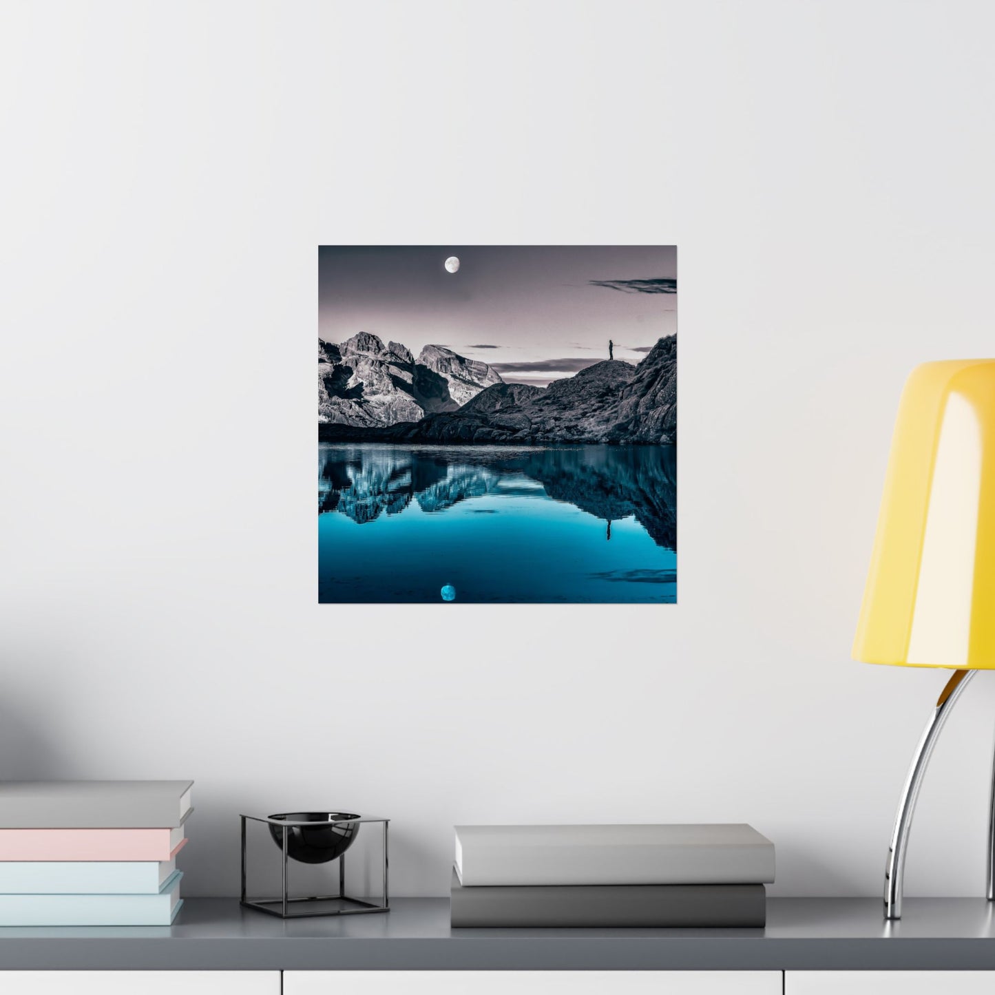 Framed landscape print of a mountainous landscape with a lake on a white wall above a shelf.