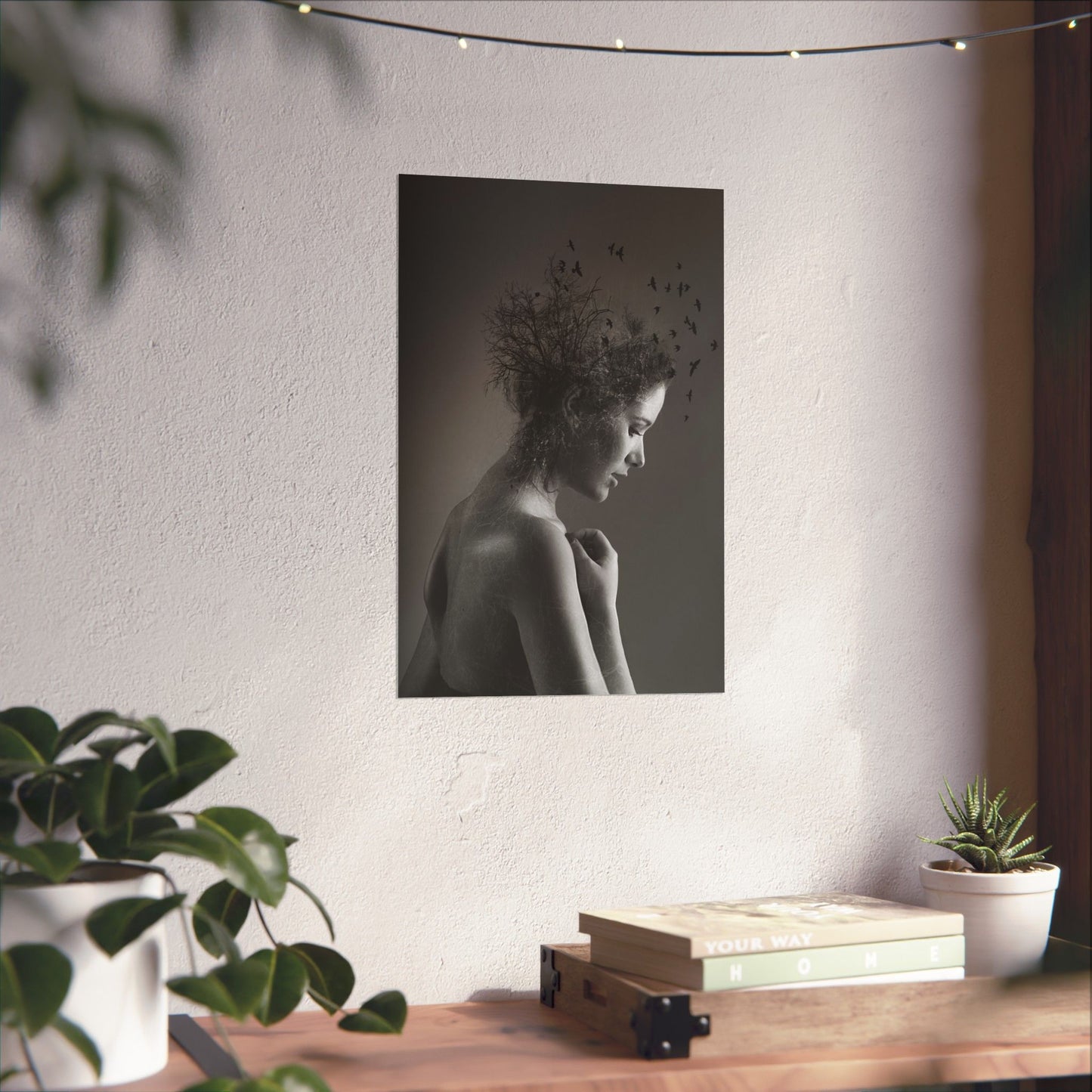 Poster of a woman with birds on her head hanging on a wall above a wooden desk with plants and books.