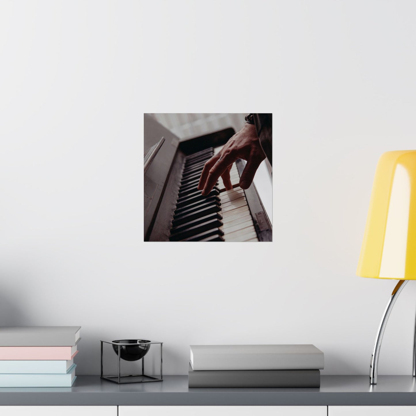 Framed picture of hands playing piano on a wall above a desk with books and a lamp.