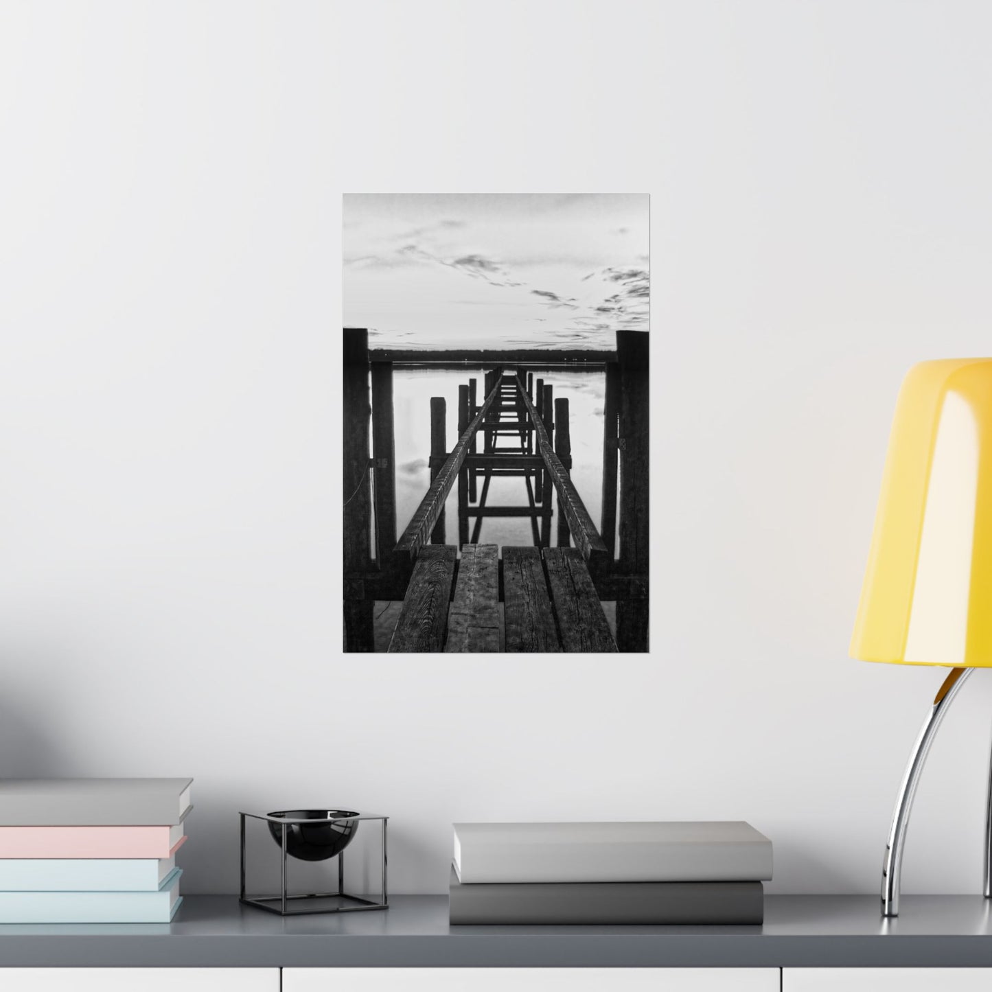 Framed black and white photo of a wooden pier on a wall above a shelf with books and decor.