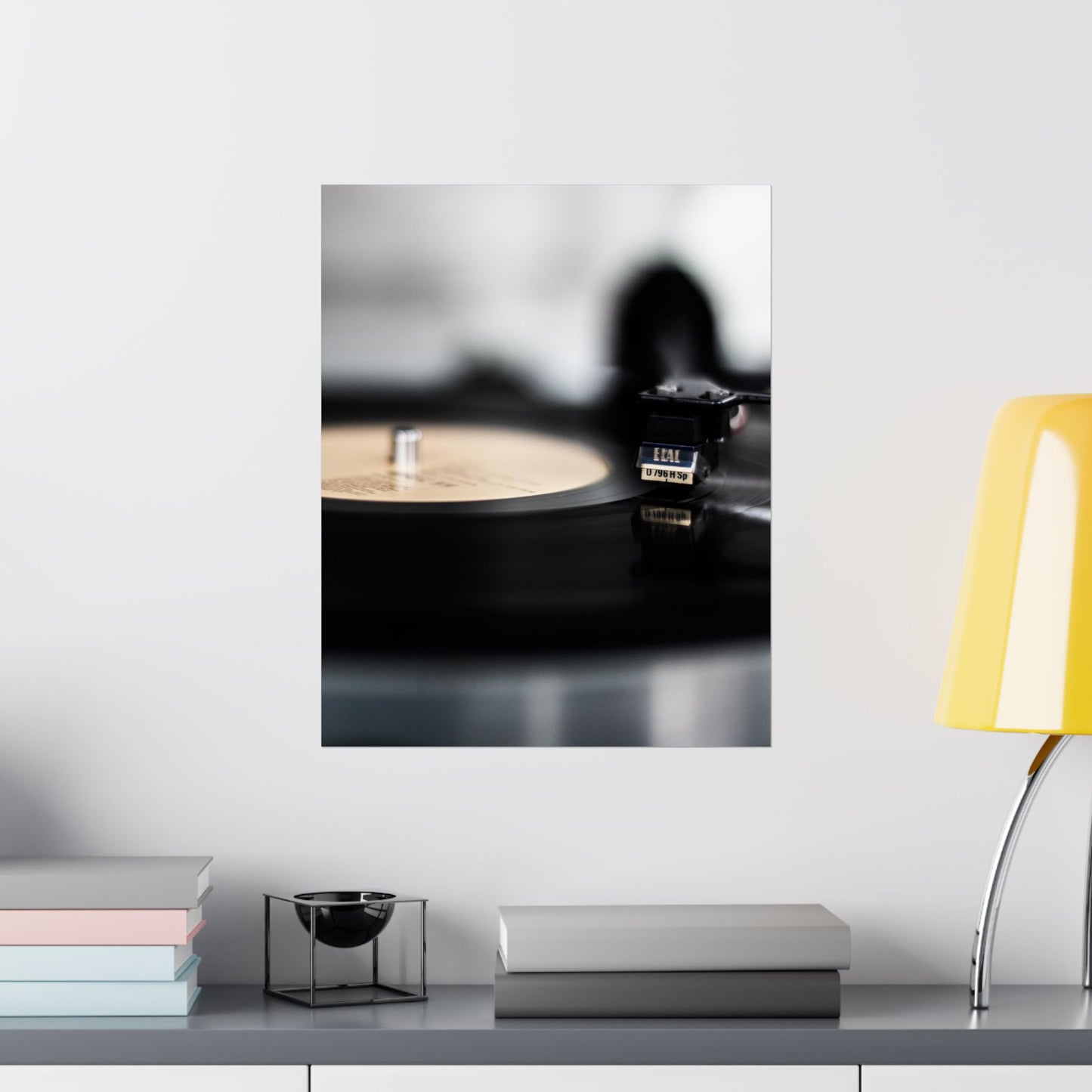 Framed artwork of a record player on a white wall with a yellow chair and books below.
