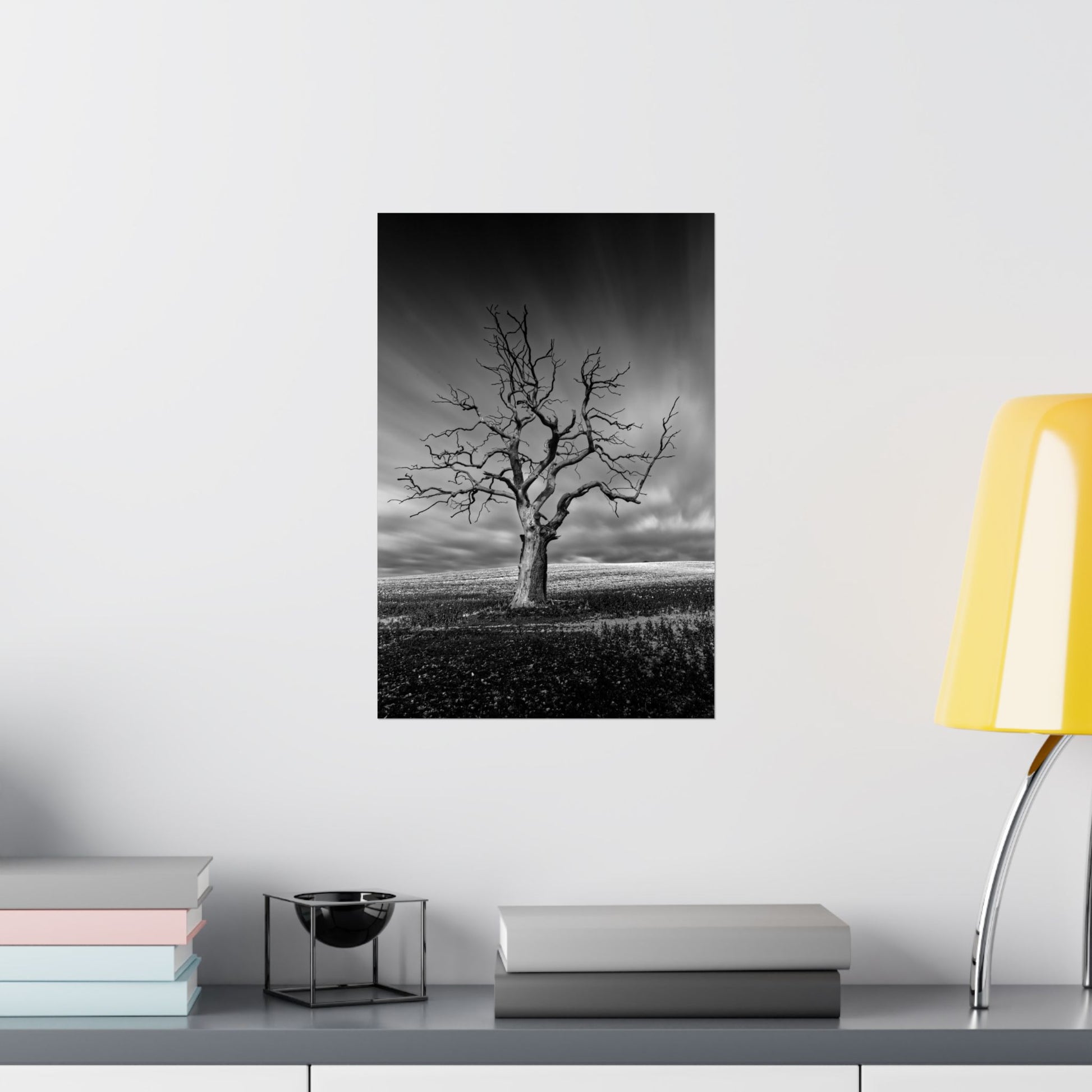 Framed black and white print of a lone tree on a wall above a shelf with books and decor.