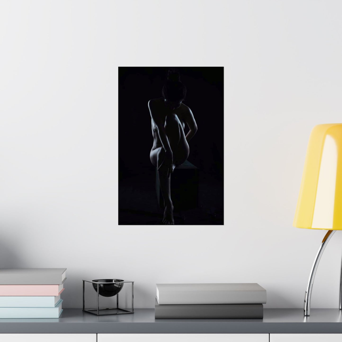 Silhouette art print of a nude figure on a wall above a shelf with books and a lamp.