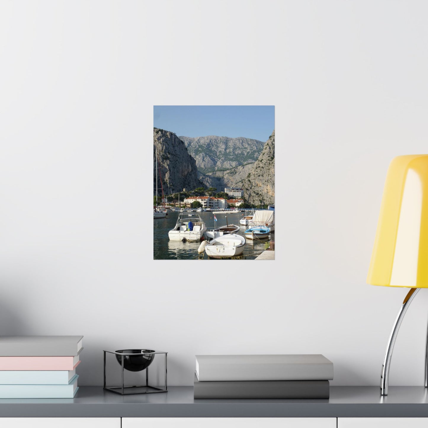 Scenic wall art of a coastal village with mountains on a white wall, next to books and a lamp.