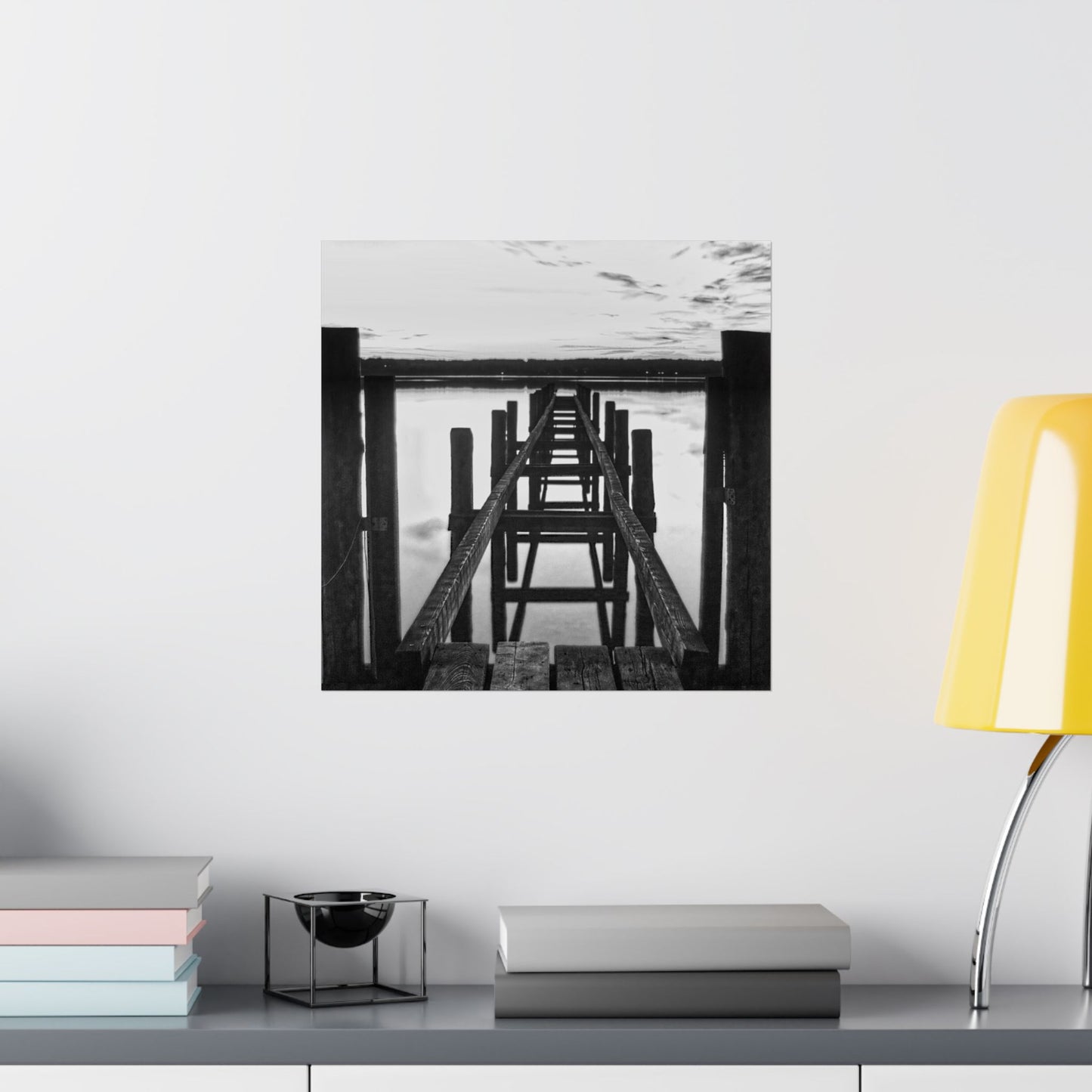Black and white photo of a pier on a wall above a shelf with books and a lamp.