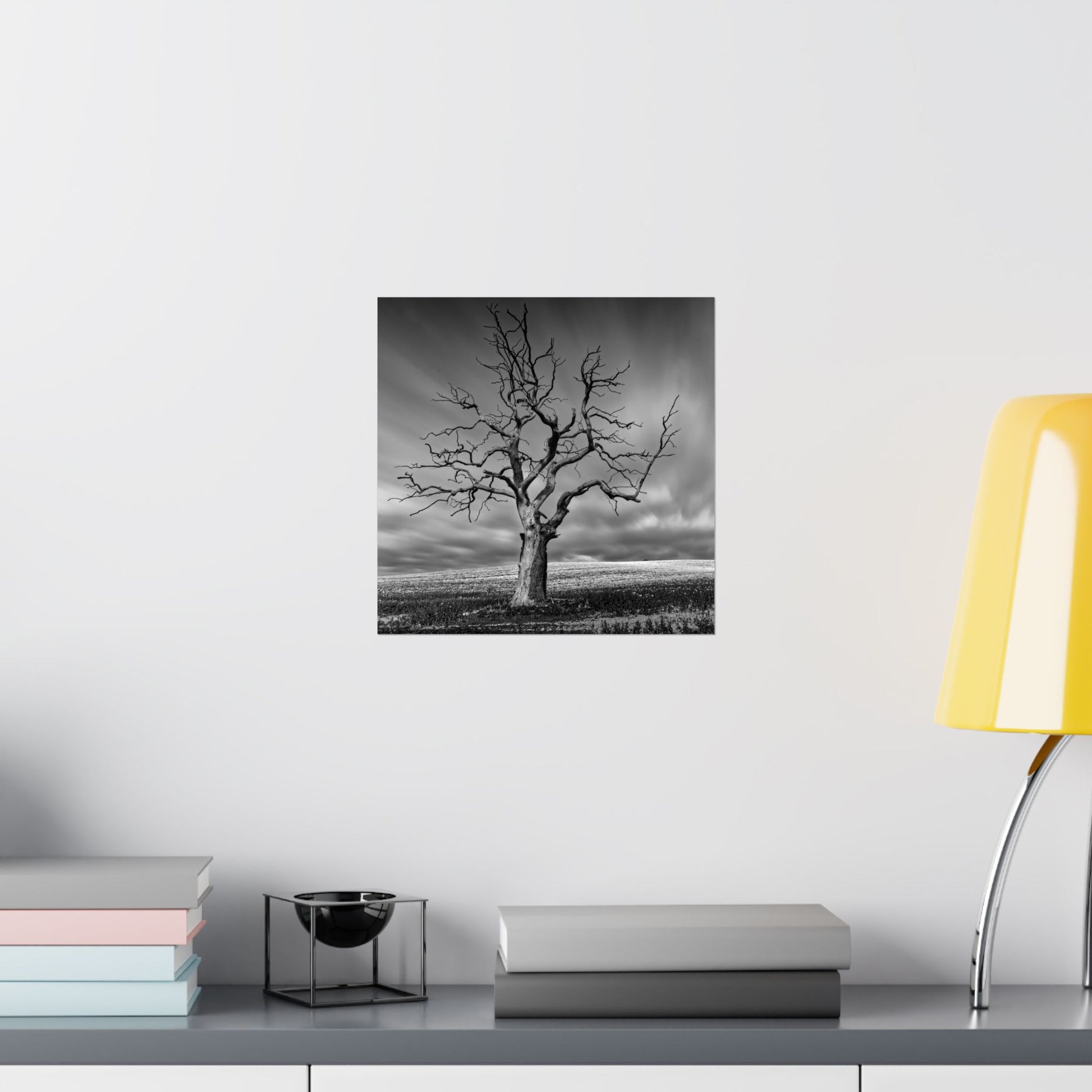 Black and white tree print on a wall above a shelf with books and a lamp.