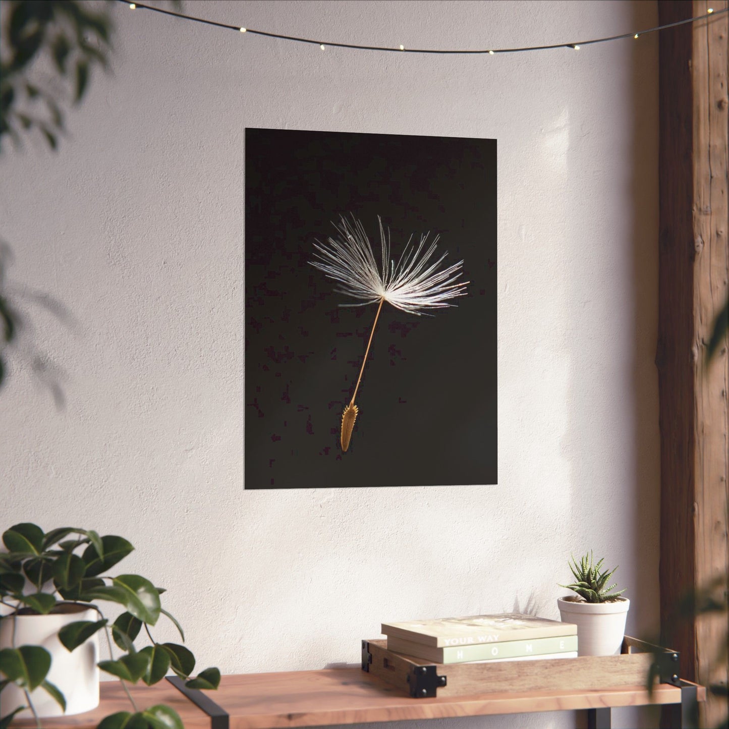 Framed artwork of a dandelion seed head on a wall with plants and books below.