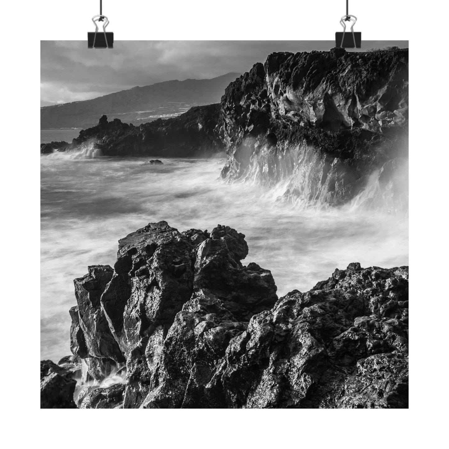 Black and white photograph of waves crashing against rocky cliffs with mountains in the background.