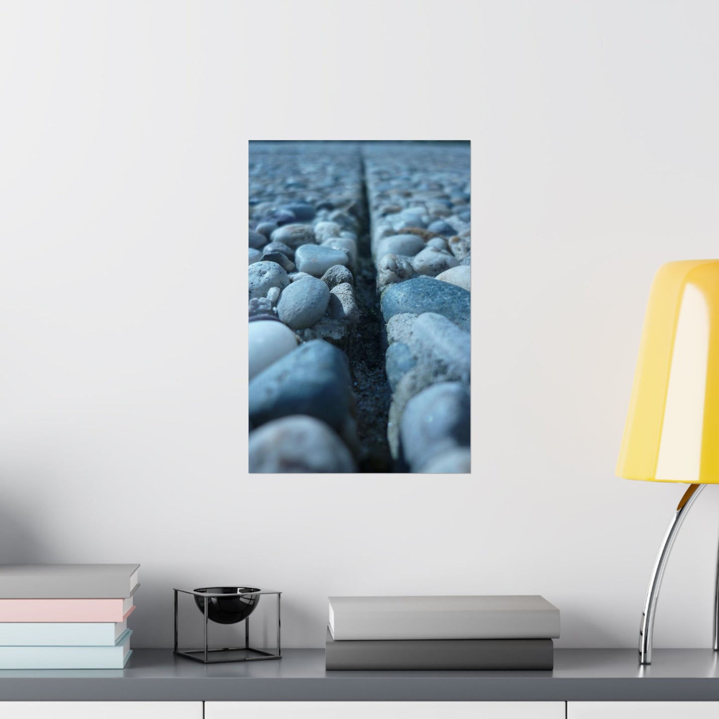 Framed artwork of pebbles on a wall above a shelf with books and a lamp.