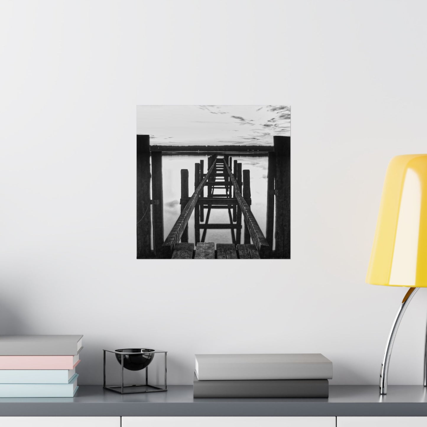 Framed black and white artwork on a wall above a shelf with books and a lamp.