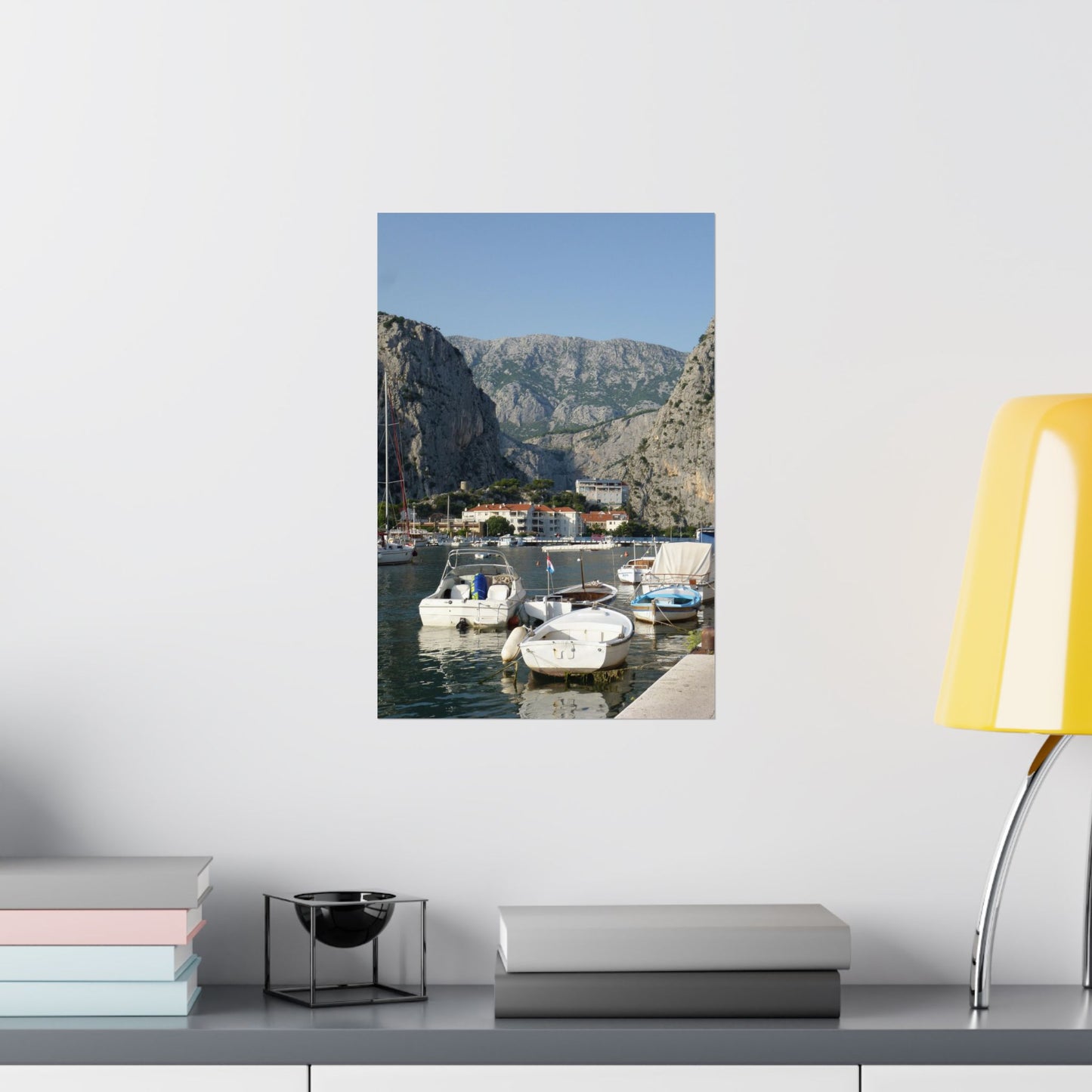 Framed photograph of a coastal scene with boats and mountains on a wall above a shelf.
