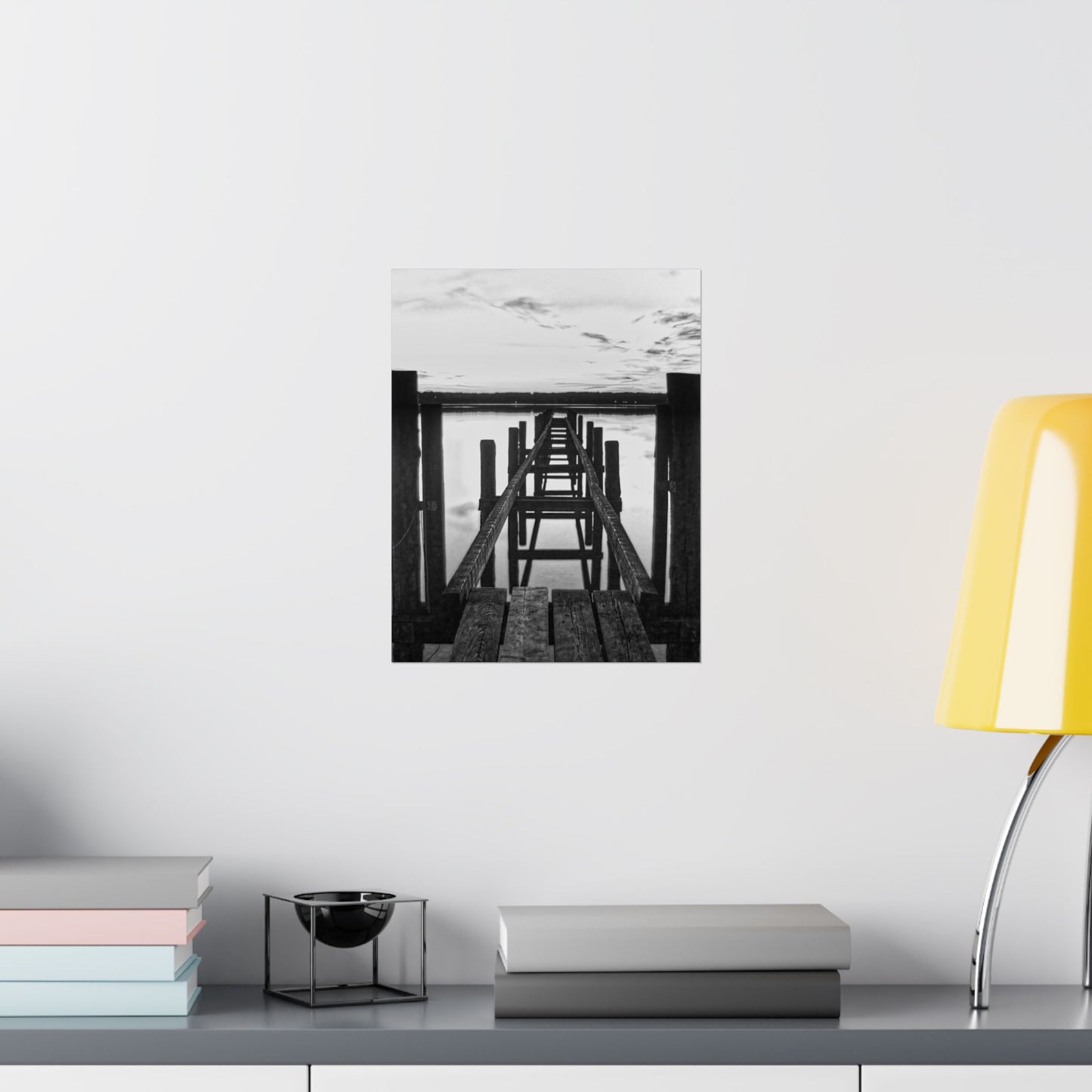 Black and white photo of a wooden pier on a wall above a shelf with books and a lamp.