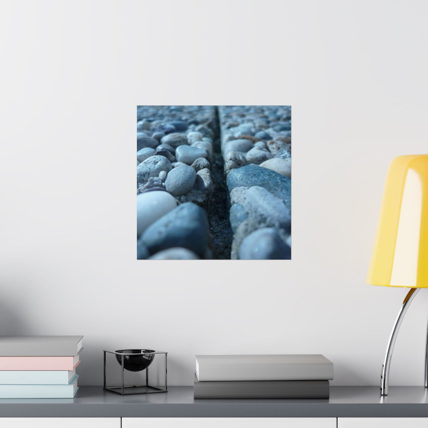 Framed artwork of pebbles on a wall above a shelf with books and a lamp.