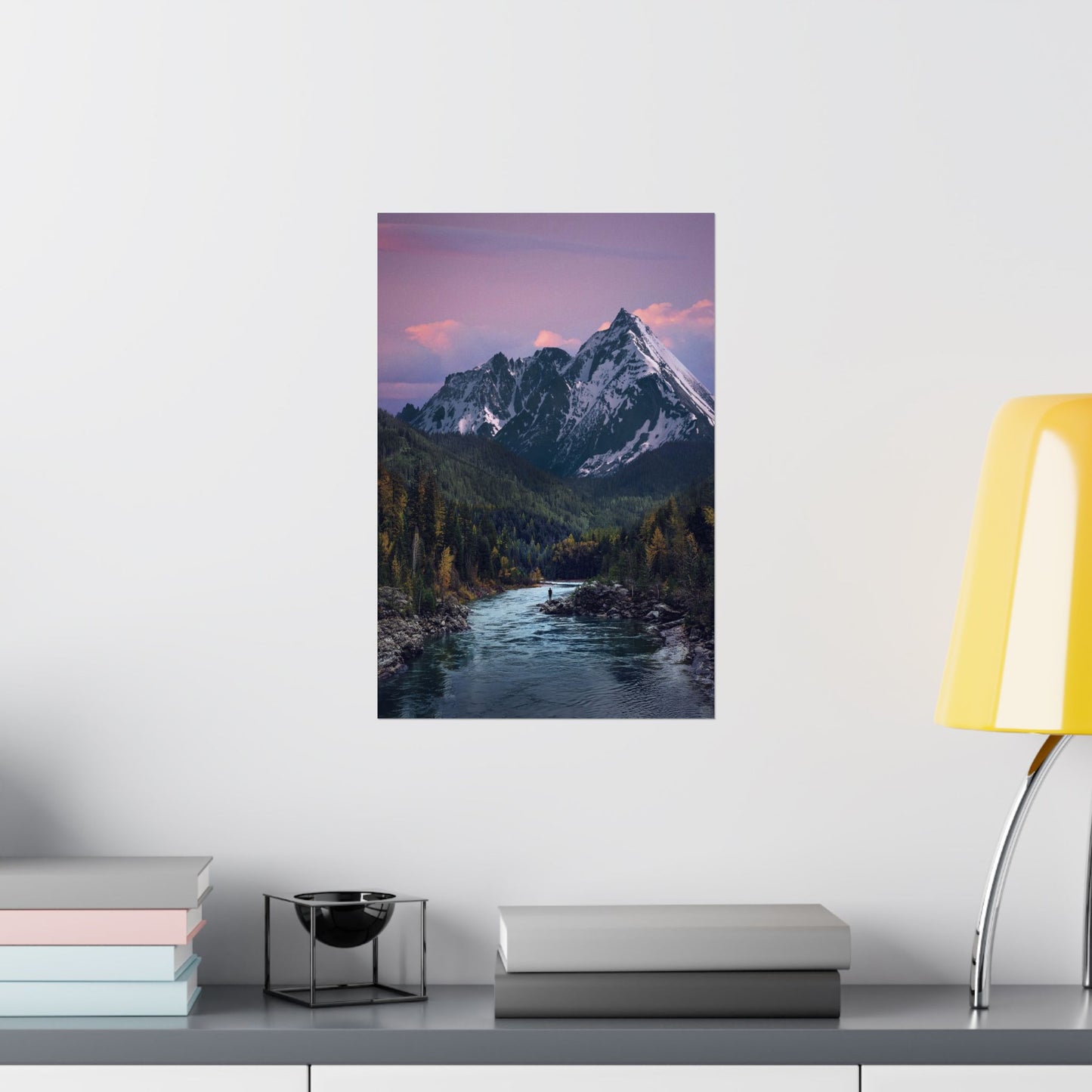Framed mountain landscape print on a wall above a desk with books and a lamp.