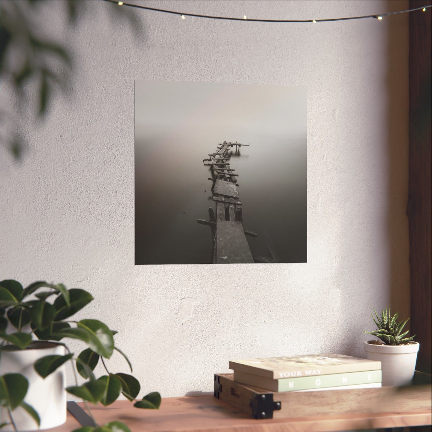 Framed artwork of a dock on a wall above a wooden table with plants and books.