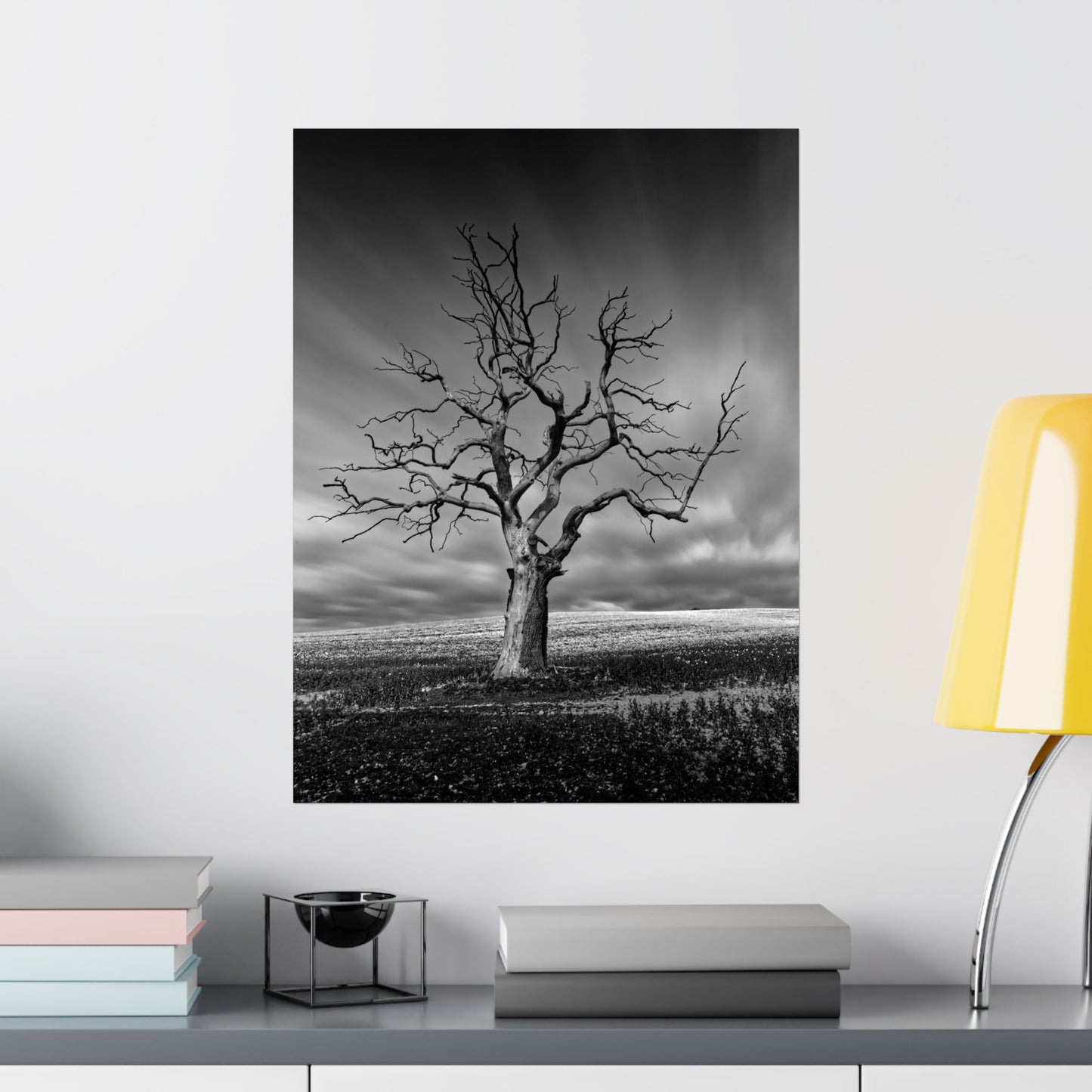 Black and white tree print on a wall above a shelf with books and a lamp.