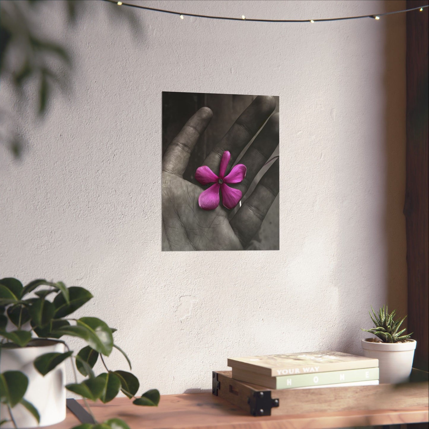 Framed artwork of a hand holding a pink flower on a wall above a wooden desk with plants.