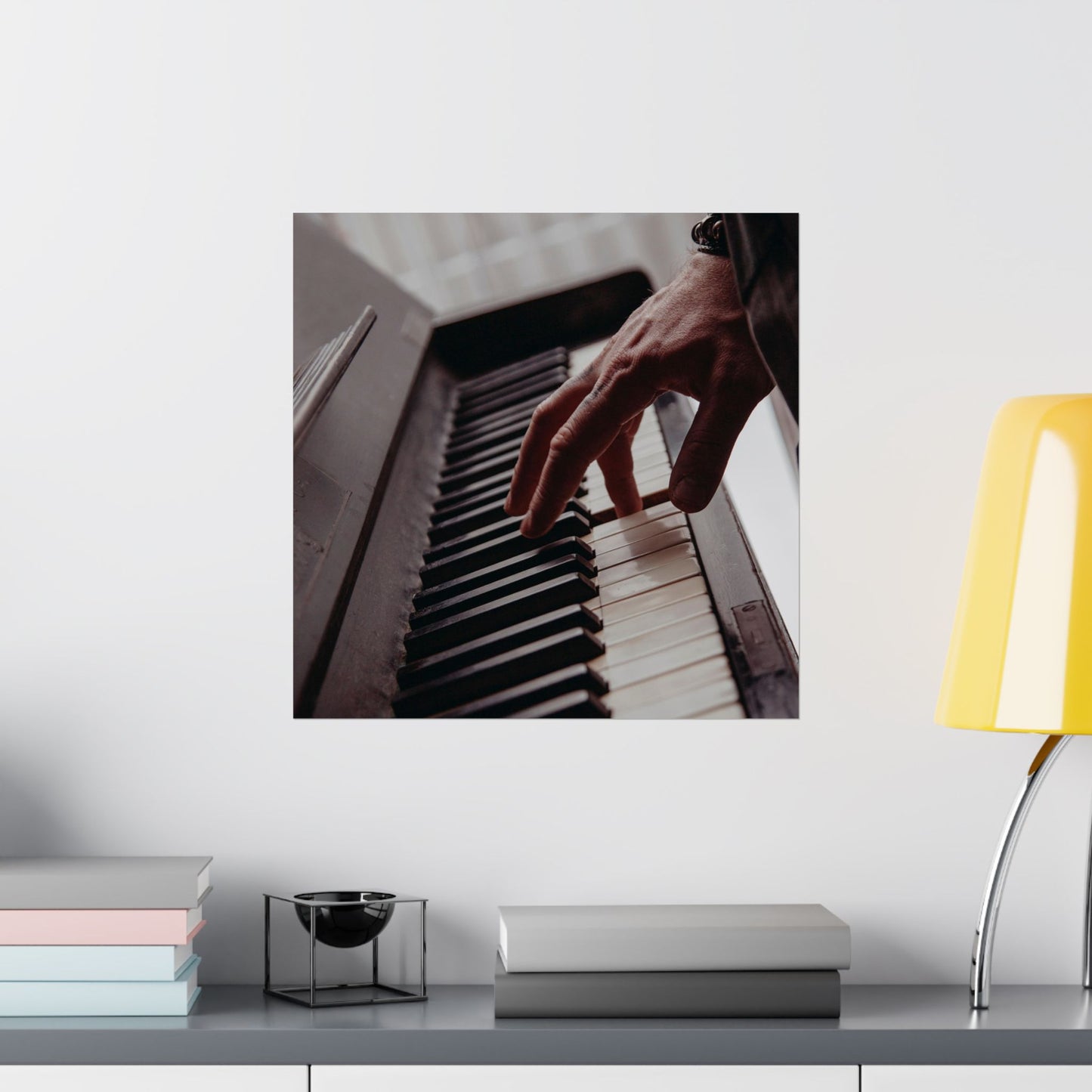 Framed picture of a hand playing piano on a shelf with books and a lamp.